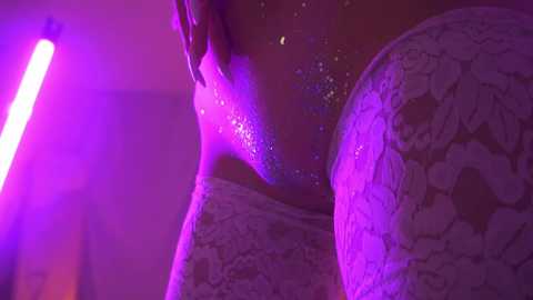 Media: A close-up video of a person with glittery, purple-tinted skin, possibly in a nightclub, highlighting their textured, illuminated face and shoulder.