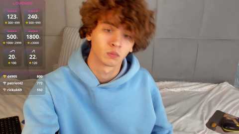 Media: Video of a young man with curly red hair, wearing a light blue hoodie, lying on a bed with white sheets and a gray padded headboard. Social media stats overlay the image.