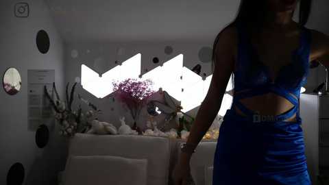 Media: Video of a woman in a blue satin dress with cutout details, holding hands with a man, standing in a dimly lit room with white walls, modern decor, and a floral arrangement in the background.