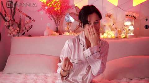 Media: Video of a woman with dark hair in a white robe, sitting on a fluffy white bed, holding a tissue, surrounded by pink and yellow lights, and decorative items.