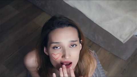 Media: Video of a young, light-skinned woman with brown hair, wearing a striped shirt, kneeling on a wooden floor, mouth open, heart drawn on her cheek, looking upward.