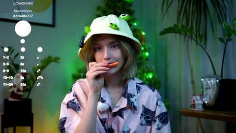 Video of a young woman with short blonde hair, wearing a white bucket hat with green frog and a pink floral shirt, holding a carrot, in a cozy, green-lit room with potted plants and a Christmas tree.