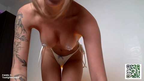 Media: Video of a topless woman with blonde hair, wearing a small bikini bottom. She has tattoos on her left arm and is leaning forward, showing her small, perky breasts.