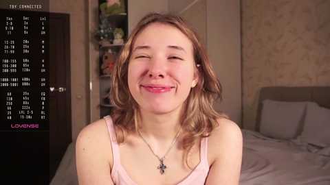 Media: Video of a smiling young woman with shoulder-length, wavy brown hair, wearing a pink tank top, in a bedroom with a cluttered bookshelf in the background.