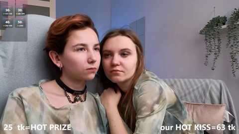 Media: Video of two women, one with short red hair and a black choker, the other with long brown hair, sitting on a couch in a room with blue walls and a potted plant.