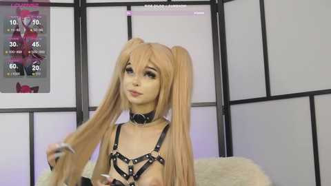 Media: Video of a light-skinned woman with long blonde pigtails, wearing a black leather harness and choker, standing in a minimalist room with white and black folding screens and a calendar.