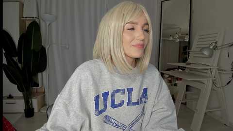 Media: A video of a young woman with straight, platinum blonde hair, wearing a grey \"UCLA\" sweatshirt, smiling, standing indoors with a white backdrop, a plant, and a desk in the background.