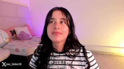 Media: Video of a young woman with long black hair, fair skin, wearing a black-and-white striped shirt, sitting on a bed with colorful pillows in a softly lit bedroom.