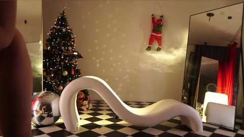 Media: Video of a modern, minimalist bedroom with a white, curvaceous chaise lounge, a decorated Christmas tree, and a large mirror reflecting festive red curtains.