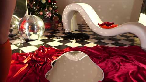 Media: A video of a festive room with a white, curvy, lit-up chair, a silver disco ball, and a red, checkered floor. A reflective, white table with a red cushion sits in the foreground.