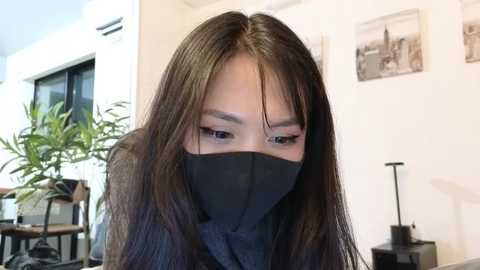 Media: Video of an Asian woman with long black hair and a black face mask, sitting indoors in a modern room with potted plants, framed photos, and a black desk lamp in the background.