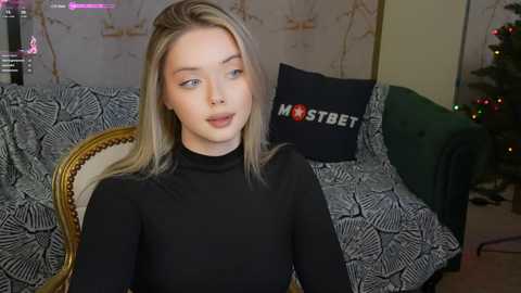 Media: Video of a young, light-skinned, blonde woman with blue eyes, wearing a black turtleneck, seated on a couch with a black and white patterned throw. Background features a green cushion, a Christmas tree, and a wall with subtle patterns.