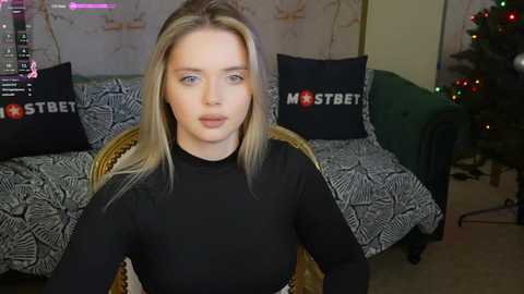 Media: Video of a young blonde woman with light skin, wearing a black long-sleeved top, seated in a room with a Christmas tree, zebra-patterned sofa, and a \"MOSTBET\" pillow.