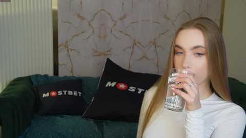 Media: Video of a young Caucasian woman with long blonde hair, wearing a white sweater, drinking water from a glass. She sits on a green couch adorned with black pillows featuring white text and a red star emblem.