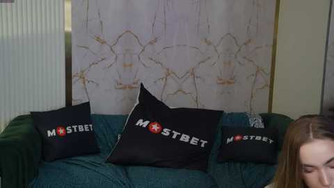 Media: Video of a woman with long brown hair, partially visible, sitting on a teal couch. The couch features three black pillows with \"MOSTBET\" logos. A white wall with gold-framed abstract art is in the background.