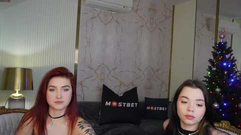 Media: Video of two women with long red and brown hair, tattoos, and dark clothes, sitting on a couch in a modern living room with a Christmas tree and decorative pillows.