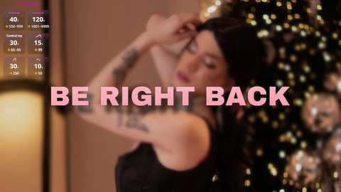 Media: Video of a woman with long dark hair and tattoos, wearing a black dress, standing in front of a Christmas tree. Text overlay reads \"BE RIGHT BACK\" in bold, pink letters.