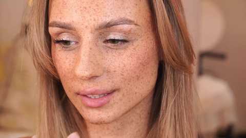 Media: Video of a fair-skinned woman with light brown hair and freckles, wearing subtle makeup, smiling slightly, indoors with a blurred background.