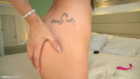 Media: A video of a nude person with light skin, holding their buttocks, revealing a tattoo on the left hip. Background includes a beige headboard and white bed linens.