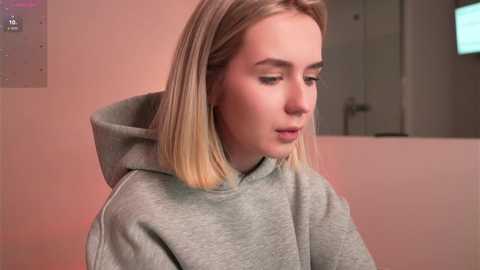 Media: Video of a young woman with shoulder-length blonde hair, fair skin, and light makeup, wearing a grey hoodie, looking contemplative against a blurred, neutral-toned background.