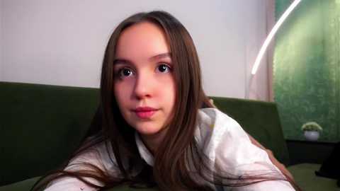 Media: Video of a young woman with long, straight brown hair, fair skin, and blue eyes, wearing a white shirt and green vest, seated on a green couch.