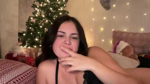 Media: A video of a smiling woman with dark hair, wearing a black top, playfully licking a wine glass in a cozy, lit bedroom with a decorated Christmas tree, a stuffed toy, and festive blankets.