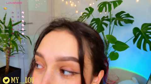 Media: Video of a young woman with fair skin, dark hair, and dark eye makeup, gazing slightly to the side. Background includes a calendar, green plants, and a glowing blue light.