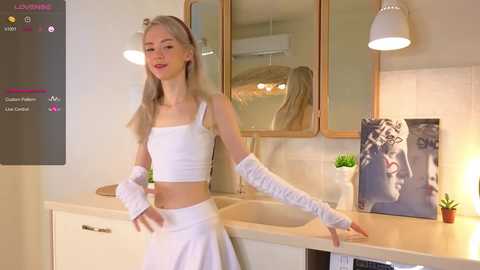 Media: Video of a slender, blonde woman in a white crop top and skirt, white arm warmers, and a headband, posing in a modern kitchen with a large mirror, potted plant, and framed picture.