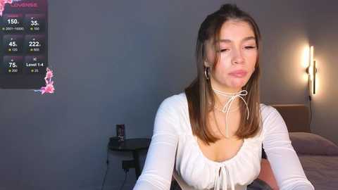 Media: Video of a young woman with light skin, straight brown hair, and a white top, sitting on a bed in a dimly lit room with a black and white wall chart and a bedside lamp.