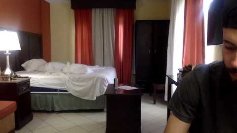 Media: Video of a dimly lit hotel room with a bed, orange and gray curtains, dark wooden furniture, and a man in a black shirt.