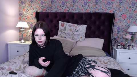 Media: Video of a young woman with pale skin, dark hair, and black clothing lying on a bed with floral-patterned wallpaper and a purple headboard, holding a smartphone.