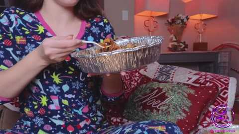 Media: Video of a woman in festive pajamas, eating popcorn from a foil tray, surrounded by Christmas decorations, including a red pillow with \"Merry\" embroidered.