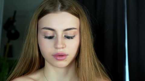 Media: Video of a young, fair-skinned woman with long, straight blonde hair, wearing natural makeup with subtle pink eyeshadow, light pink lipstick, and well-groomed eyebrows. Background shows a dark curtain and a guitar partially visible.