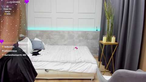 Media: Video of a modern, minimalist bedroom with a gray headboard, white bedding, and a gray chair; a wooden side table with a potted plant.