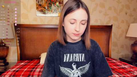 Media: Video of a young Caucasian woman with straight, shoulder-length brown hair, wearing a dark \"Avenged Sevenfold\" sweatshirt, sitting on a bed with a red and black plaid blanket.