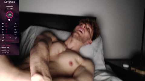 Media: A video of a shirtless, muscular, fair-skinned man with red hair, lying in bed, moaning with eyes closed, surrounded by gray sheets and pillows, with a digital screen overlay showing \"Lowenbake\" and a stats menu.