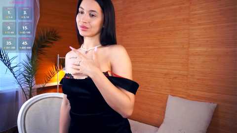 Media: Video of a slender, fair-skinned woman with long black hair in a black satin dress, holding a glass, in a modern, warmly lit room with wooden paneling and white cushions.