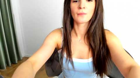 Media: Video of a young woman with long, straight brown hair, fair skin, wearing a light blue tank top, sitting indoors, with a green curtain in the background.