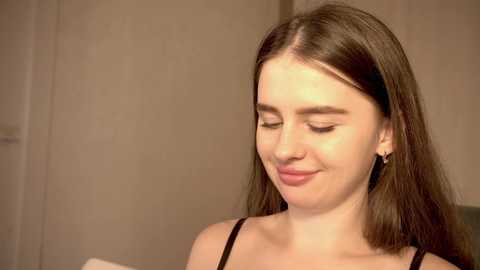 Media: Video of a young Caucasian woman with long brown hair, wearing a black spaghetti-strap top, smiling gently with closed eyes. Background features a beige wall and indistinct objects.