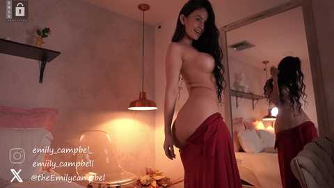 Video of a topless woman with long dark hair, wearing a red skirt, in a dimly lit, cozy room with a bed, plants, and a mirror.