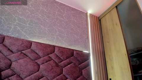 Media: Video of a modern bedroom corner featuring a plush, diamond-patterned maroon headboard against a textured, silver wall. A wooden wardrobe with vertical slats and a soft, warm light accentuating the corner.