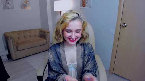 Media: Video of a smiling woman with blonde, shoulder-length hair, wearing a patterned cardigan, seated in a beige armchair in a modern, light-colored room with a beige couch and a white lamp.