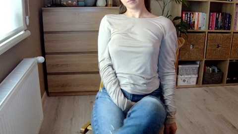 Media: Video of a fair-skinned woman with medium-length brown hair, seated on a chair in a bright room. She wears a light grey long-sleeve top and blue jeans. Wooden dresser and bookshelf in background.