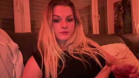 Media: Video of a blonde woman with long hair, wearing a black shirt, sitting on a couch in a dimly lit room with orange lighting, holding a glass of red wine.