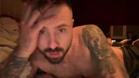 Media: Video of a shirtless, tattooed man with short hair, resting his head on his hand, looking pensive. He has a beard and is in a dimly lit bedroom with dark wooden furniture.