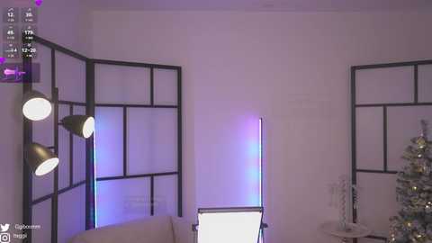 Media: Video of a minimalist, modern living room with white walls, black-framed glass panels, and a Christmas tree in the background. A large, adjustable light stand with multiple heads casts a soft, blue glow.