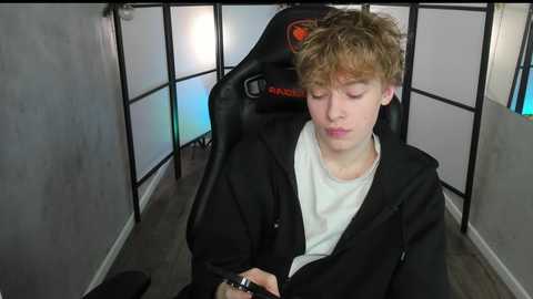 Media: Video of a young man with curly hair, wearing a black hoodie and white shirt, seated in a black gaming chair, with a \"Razer\" logo, in a dimly lit room with gray walls and a colorful neon light.