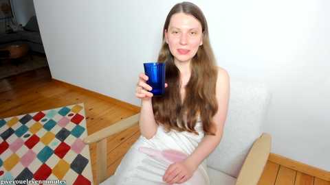 Media: Video of a young Caucasian woman with long brown hair, wearing a white dress, sitting on a wooden chair with a colorful rug in a minimalist room.