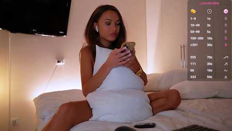 Media: Video of a light-skinned, brunette woman in a white dress, sitting on a bed, holding a phone, surrounded by white pillows and a flat-screen TV.