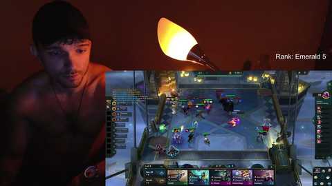 Media: A video with a man in a black beanie, shirtless, standing in a dimly lit room. The background displays a computer screen playing a video game, showing an arena with colorful characters and bright lights.